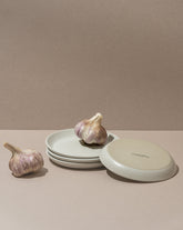 A white stoneware flat side plate with a matte finish, displayed alongside garlic bulbs. The plate is placed against a neutral background.